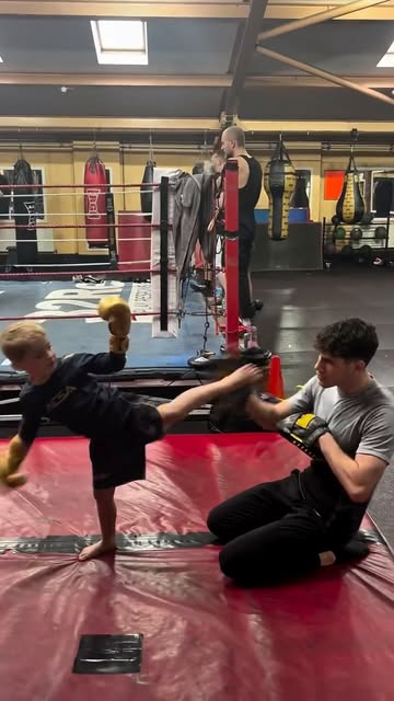 Tom Brennan kneeling on the mat holding a kick pad while a young student throws a head kick.