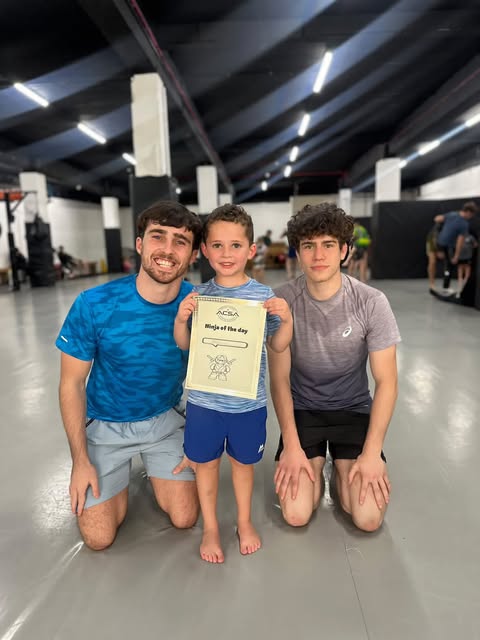 Tom Brennan and a fellow coach kneeling with a young student holding a ninja of the day certificate.